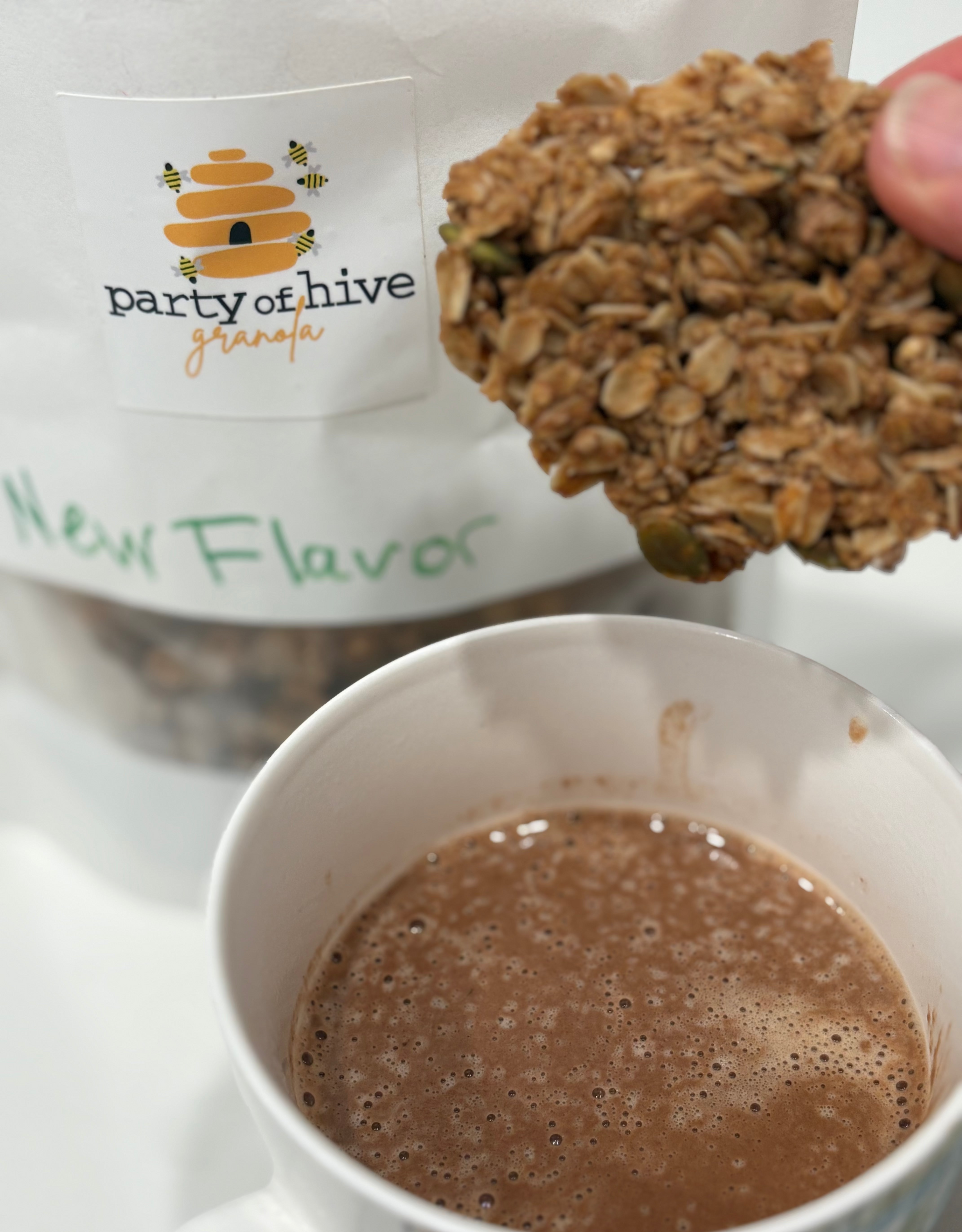 Granola cookie held above a cup of hot chocolate with 'party of hive' granola packaging in the background.